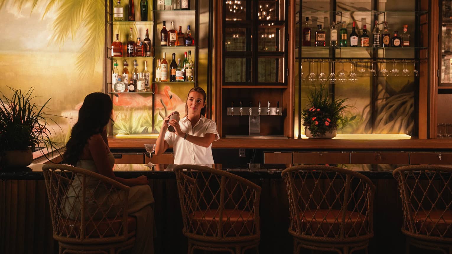 Bartender mixes a drink for a guest at a bar with a mural of a beach on the wall