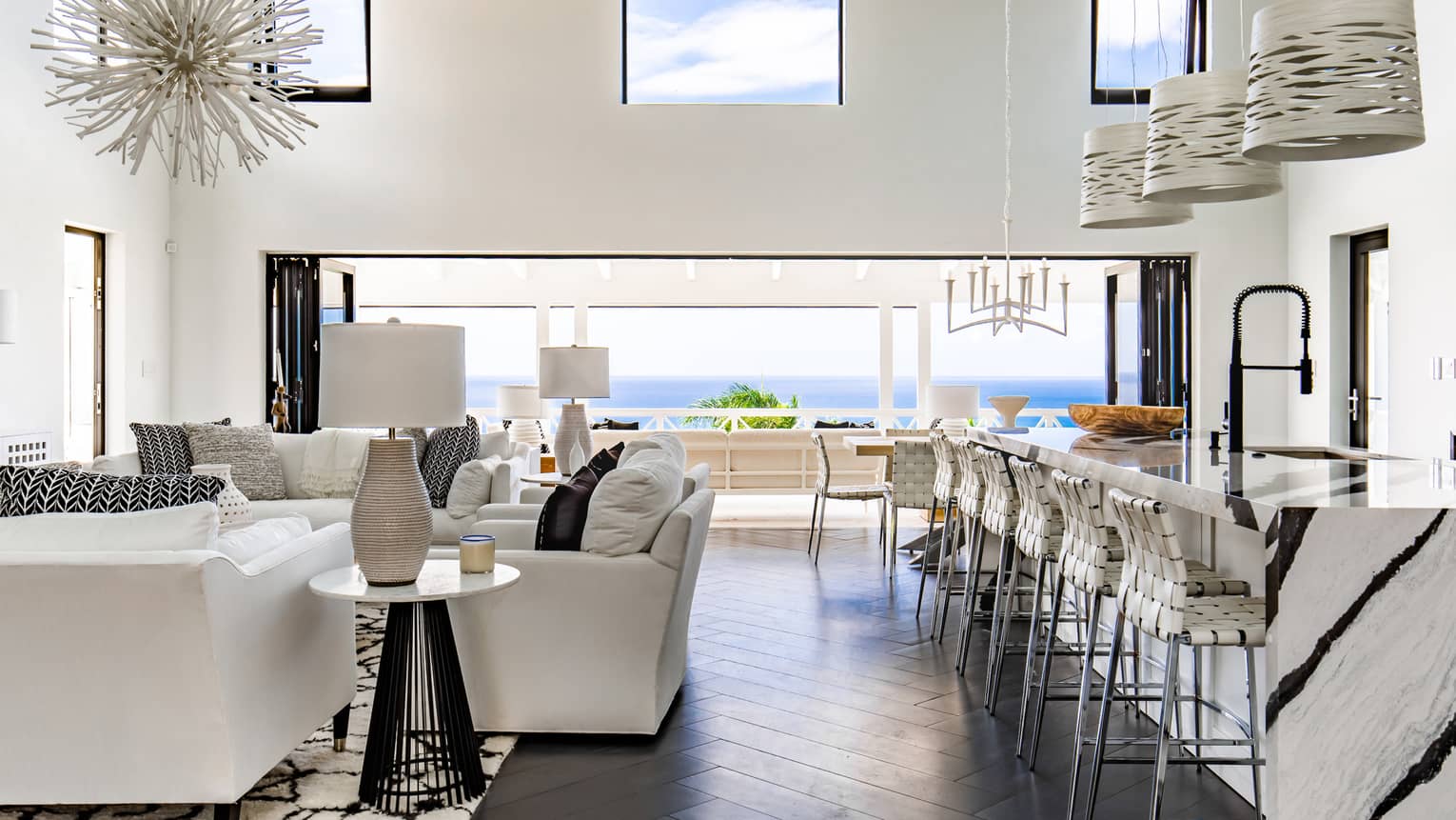 Large common area with long marble kitchen island and bar chairs, white living room furniture, sea view