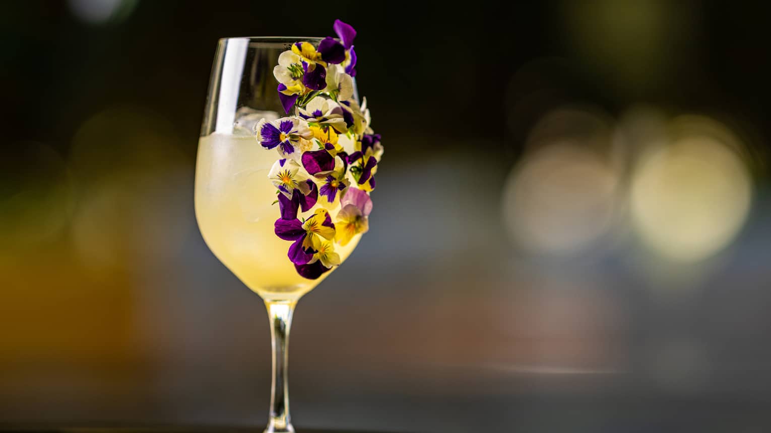 A wine glass with a yellow drink and flowers on the side of the glass