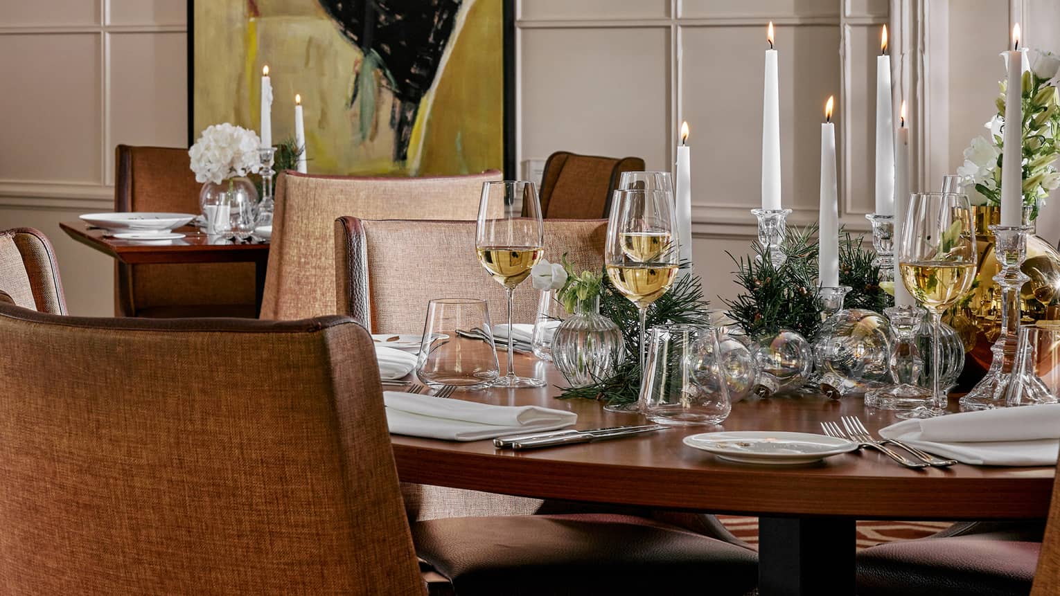 Elegantly set holiday table with greenery, lit white taper candles, glasses of white wine and white porcelain dishes