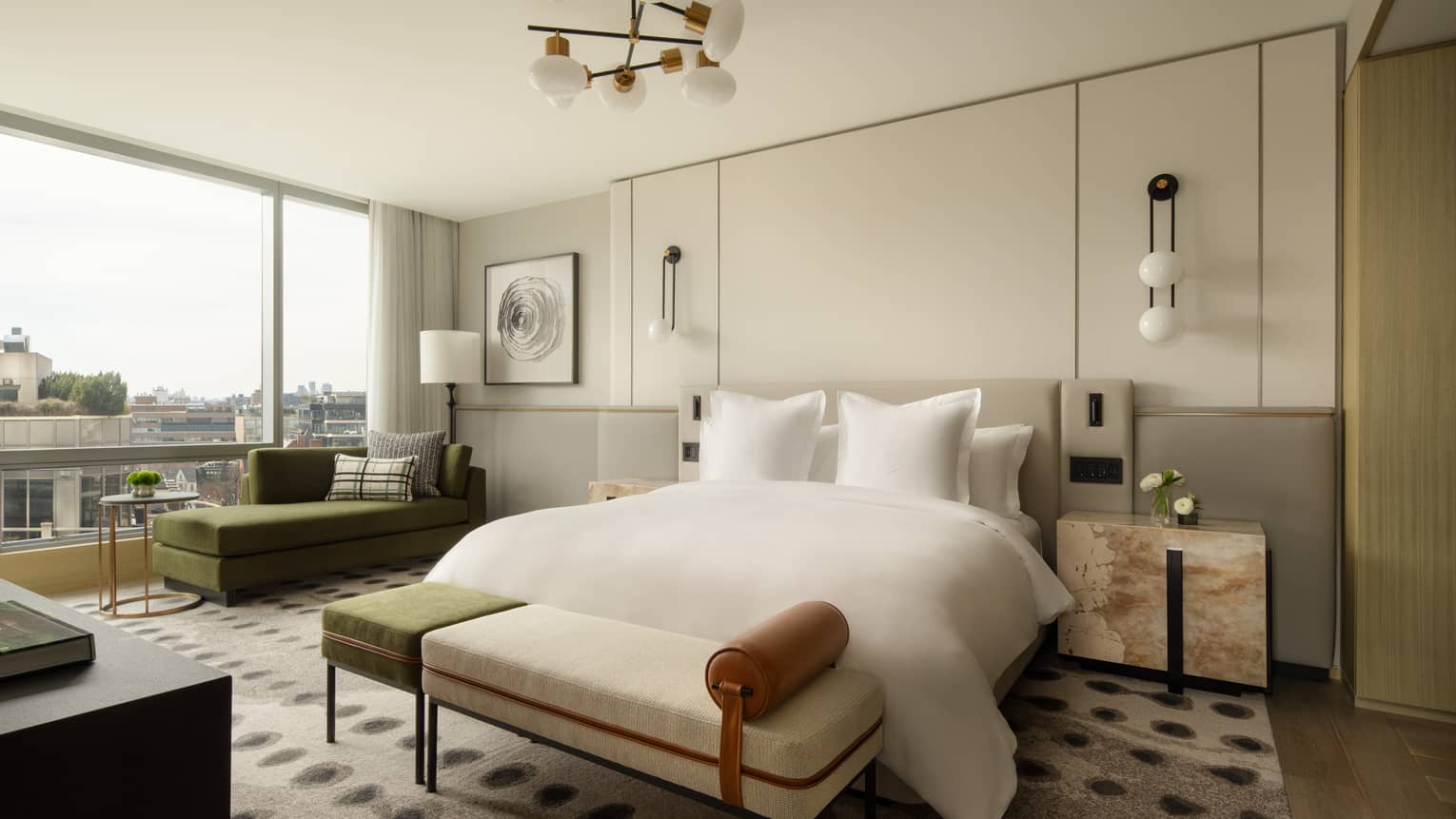 Luxury hotel suite bedroom with king bed, modern side tables, bench, green chaise lounge by the floor-to-ceiling window and patterned rug