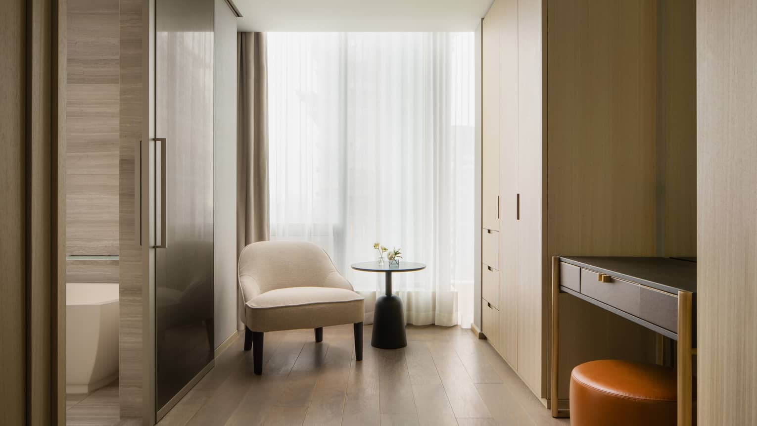 Dressing area with a beige armchair, round black side table with a flower arrangement, built-in wardrobes and a vanity desk with an orange ottoman