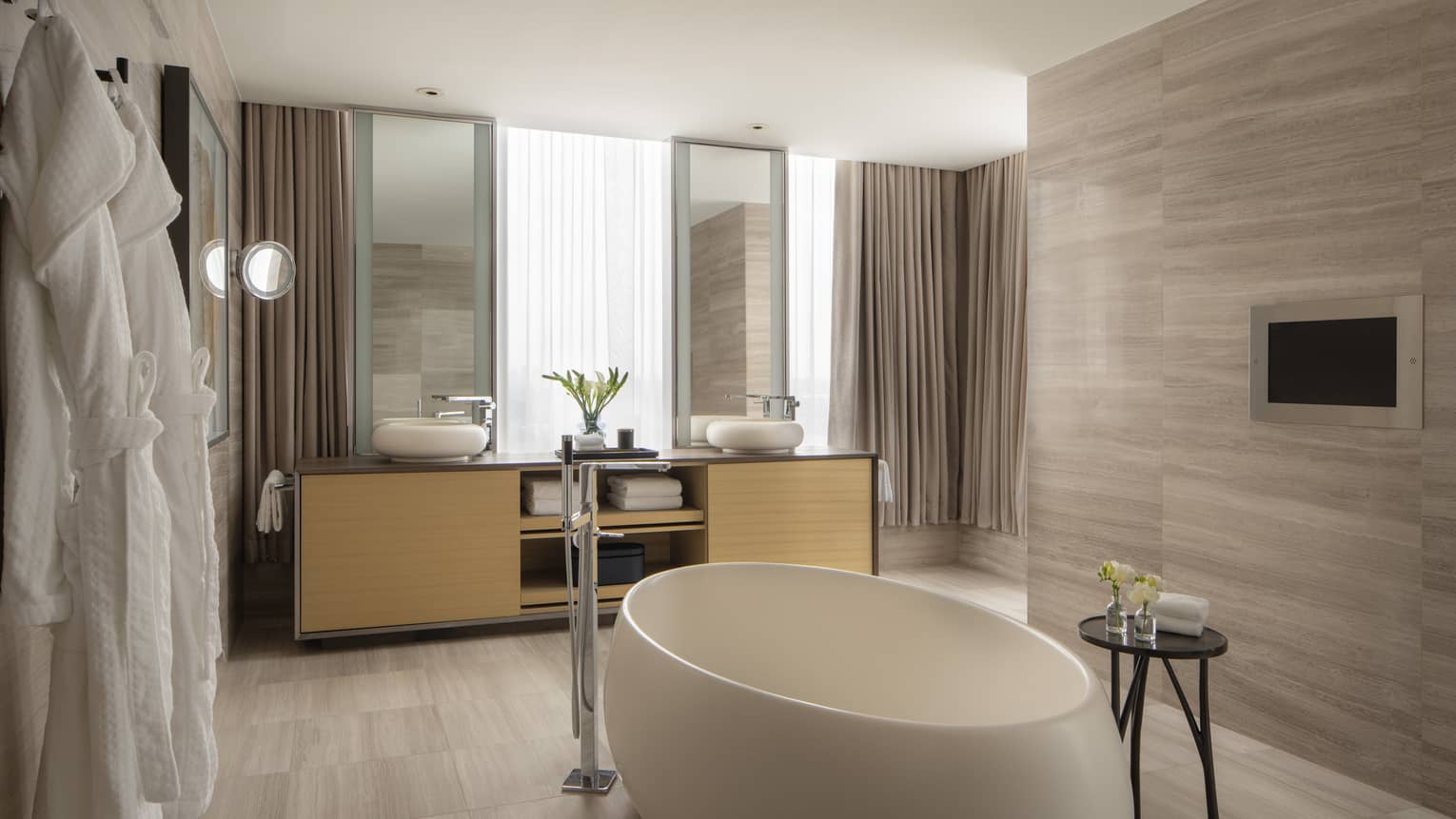 Luxury suite bathroom with a freestanding oval tub, double vanity, light wood finishes and large windows letting in natural light