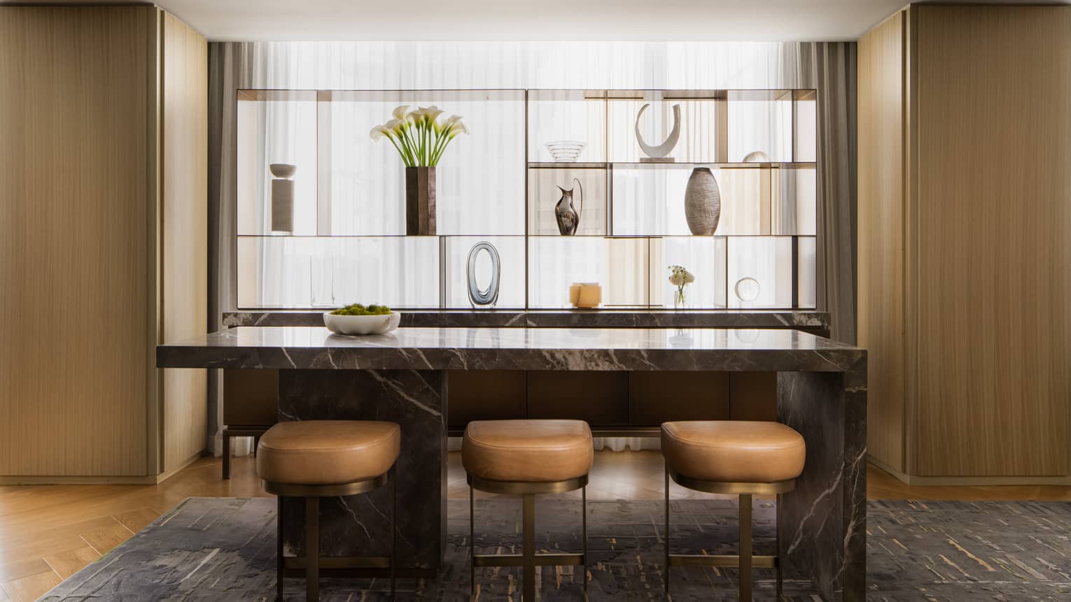 Luxury suite bar area with marble island, three leather stools, and decorative display shelf