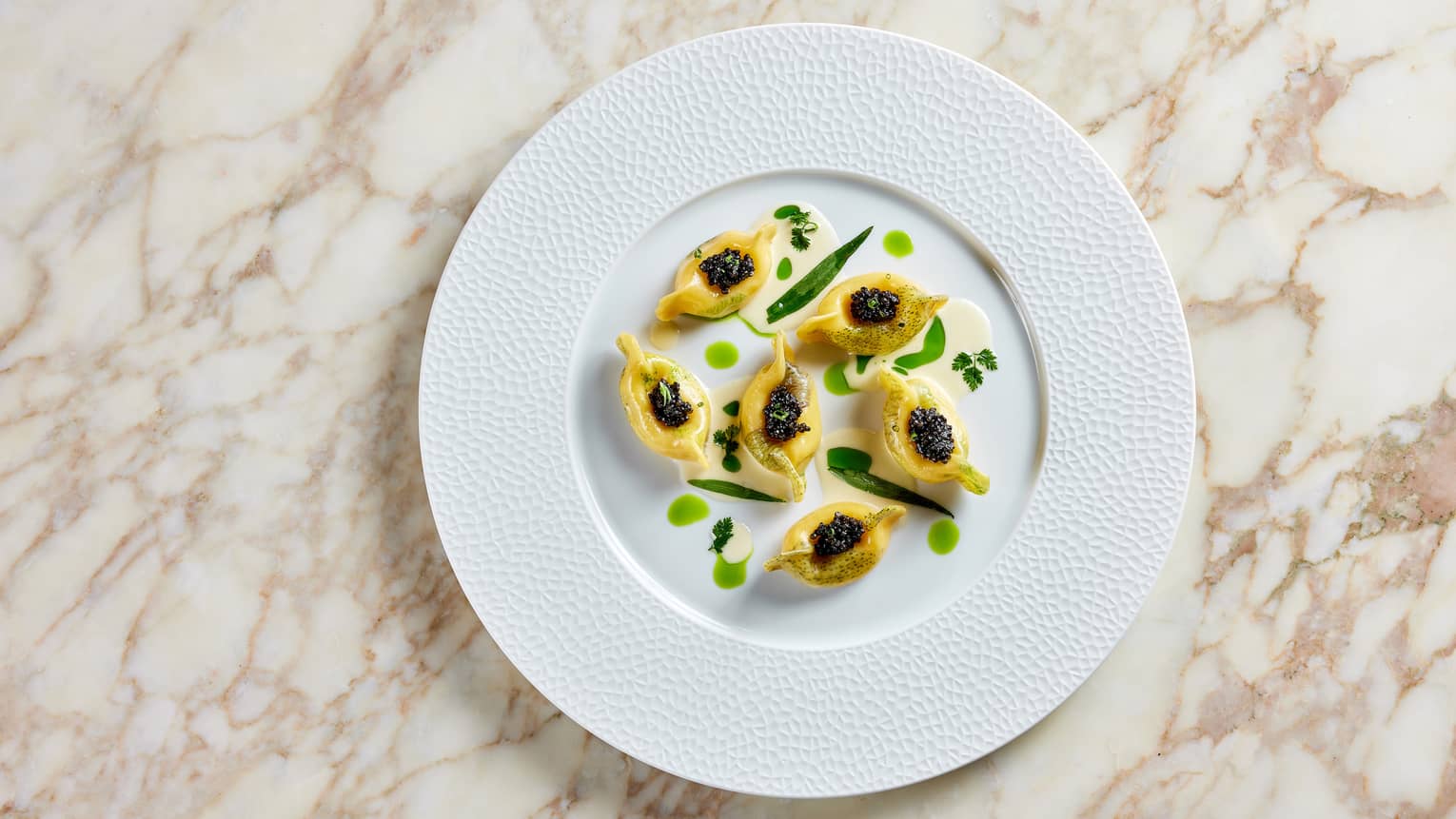 On a round white plate, six blinis garnished with black caviar and herbs over a creamy sauce, set on a marble surface.