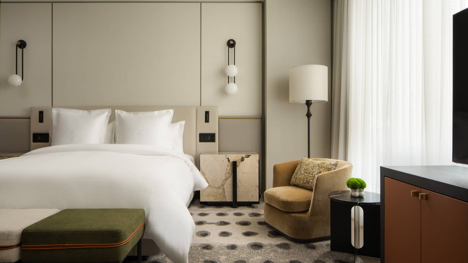 Hotel suite bedroom with a king bed, nightstand, armchair, floor lamp and TV console against floor-to-ceiling windows