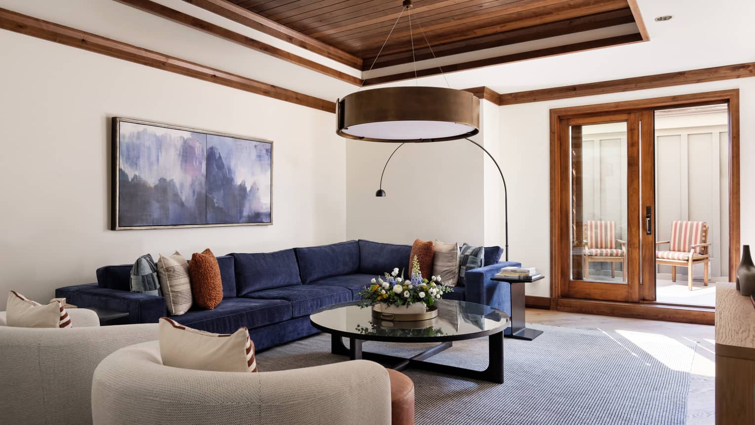 Living room with a navy sectional sofa, round coffee table with floral centrepiece, two armchairs and wood-framed glass doors opening to a patio