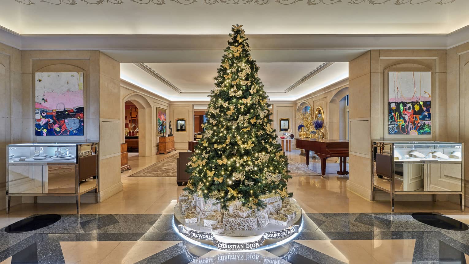 Luxurious hotel lobby, featuring a large elegantly decorated Christmas tree with gold ornaments and lights, surrounded by Christian Dior gift boxes and modern artwork on the walls.