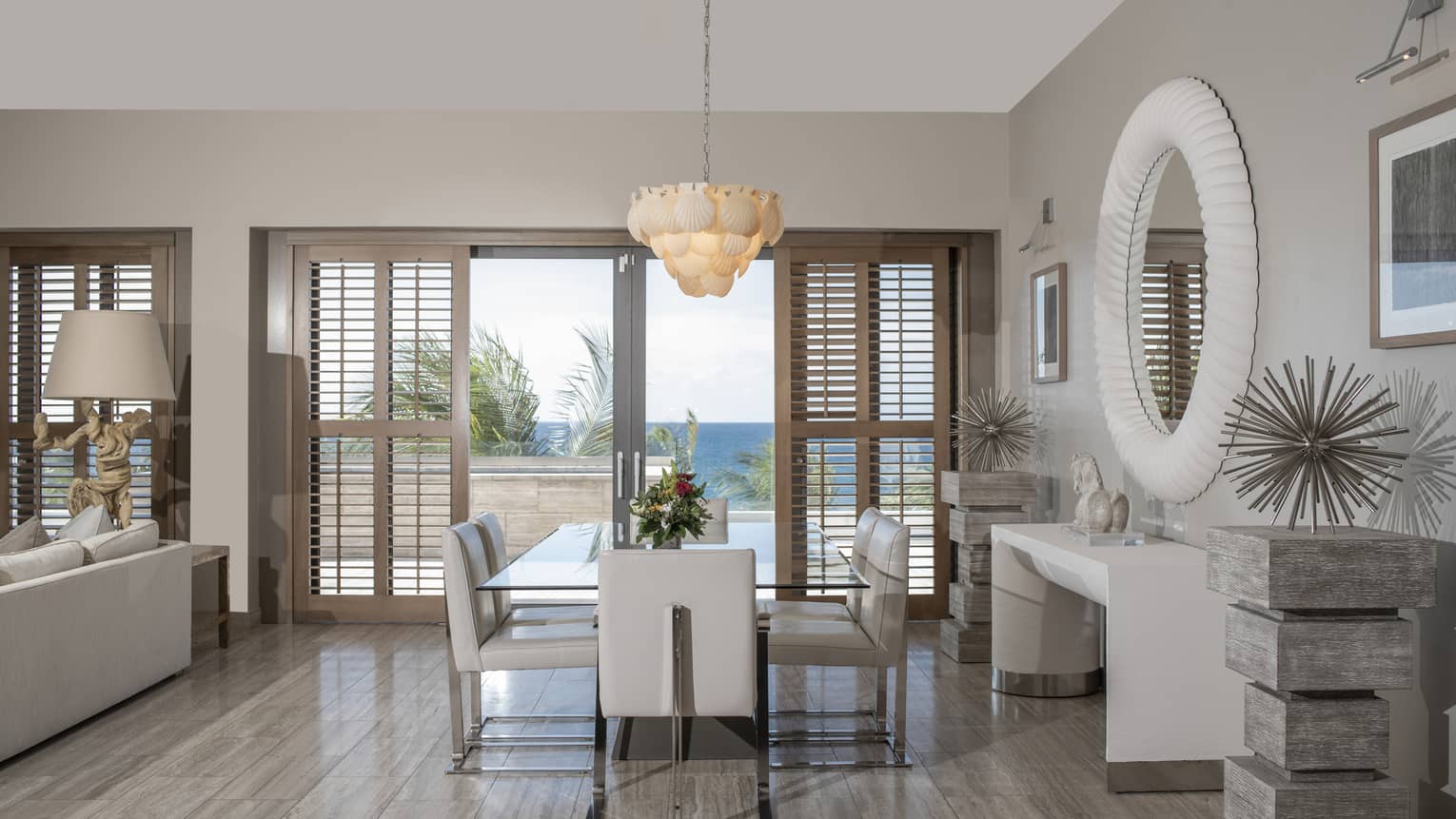 Luxury resort penthouse dining area with a glass table, white leather chairs, coastal decor and large windows with wooden shutters offering an ocean view