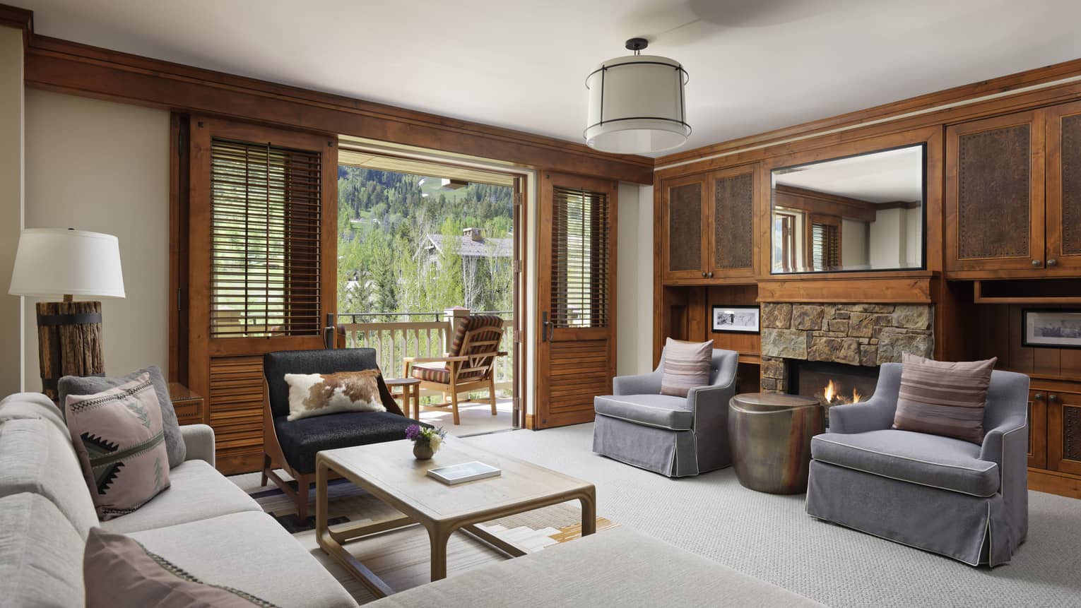 Resort living area with a stone fireplace, light grey sofa, two armchairs, rustic wooden cabinetry and a private balcony with mountain view