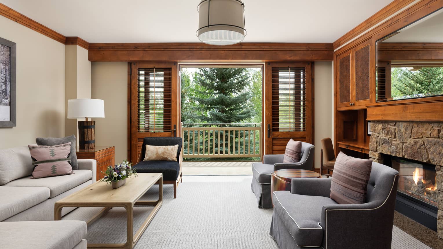 Luxury resort living room with a light grey sectional sofa, armchairs, coffee table, stone fireplace, and walk-out balcony with a view of evergreen trees