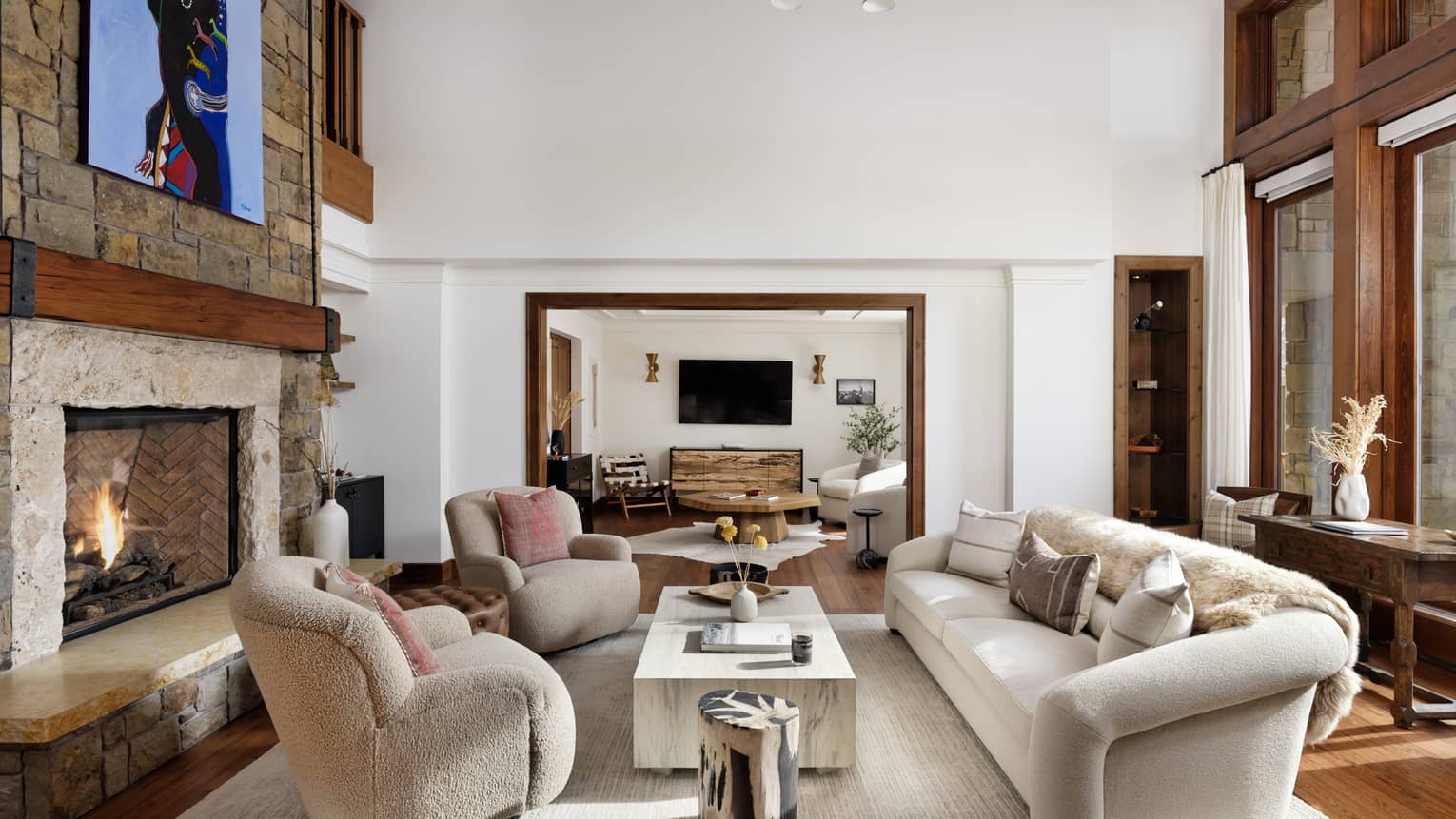 Mountain residence living room with a stone fireplace, sofa and armchairs around a coffee table, leading into a connecting seating area with a TV on the wall