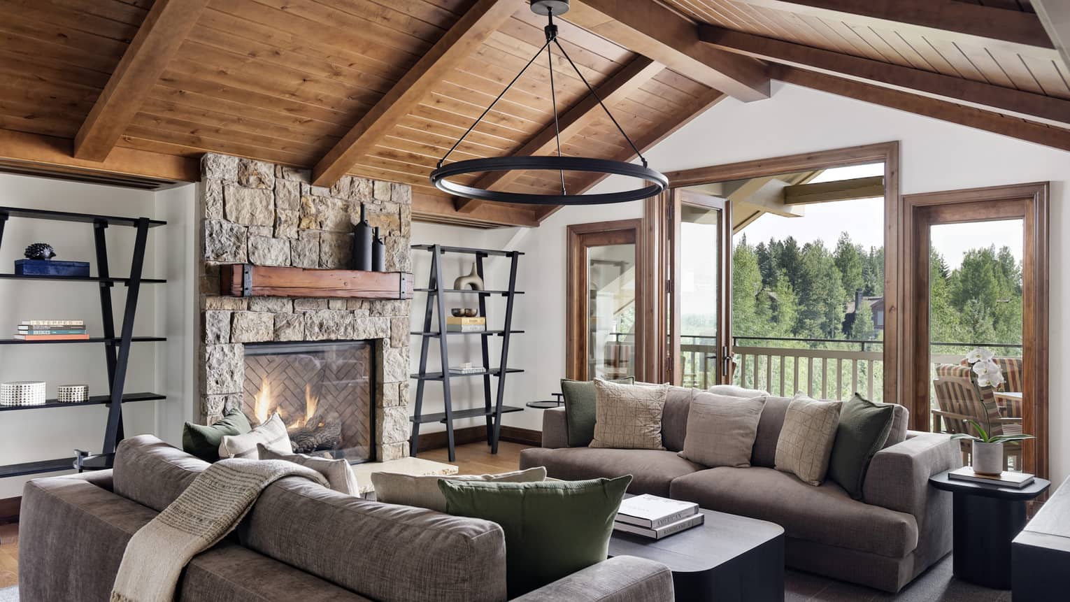 Granite residence living room with vaulted wood ceiling, stone fireplace, grey sofas and a circular chandelier, opening to a balcony with forest view