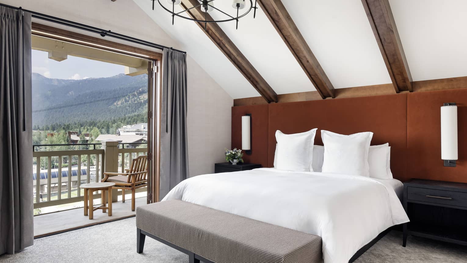 Bedroom with vaulted wood-beam ceiling, rust-coloured headboard, king bed with white linens, grey bench and open balcony with mountain views