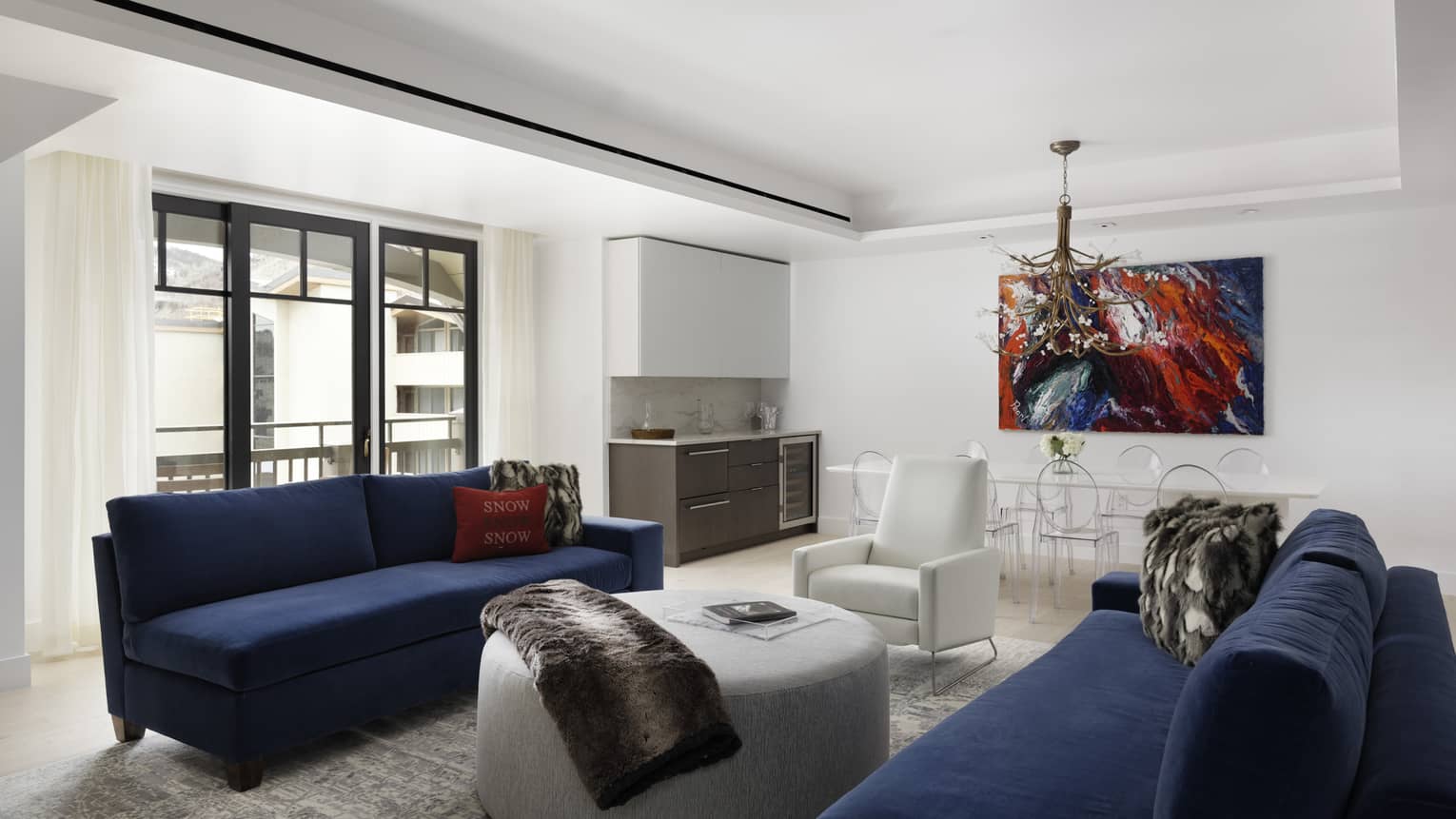 Living room with white walls and ceiling, two dark blue sofas, light grey round ottoman, artwork, windows