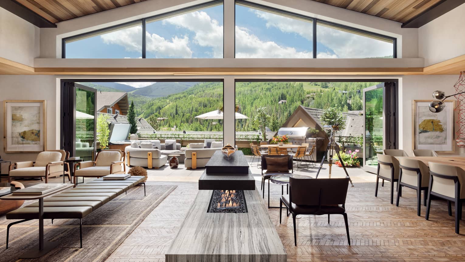 Luxurious mountain villa interior with an open-plan living area, modern fireplace, and expansive glass doors leading to an outdoor terrace with panoramic forest views.