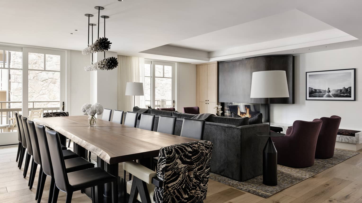 A large wooden dining table with 14 chairs set next to a living room with sofa, two arm chairs and fireplace