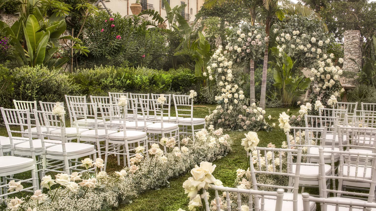 A garden wedding setup featuring rows of white chairs along a floral aisle leading to a flower arch