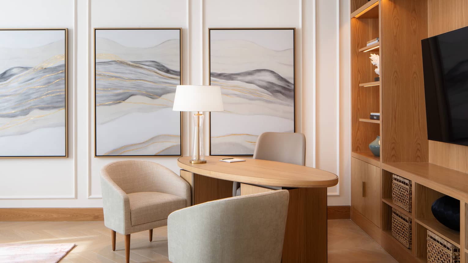 Modern home office with a curved wooden desk, neutral chairs, abstract wall art and built-in shelving with decorative accents and storage baskets.