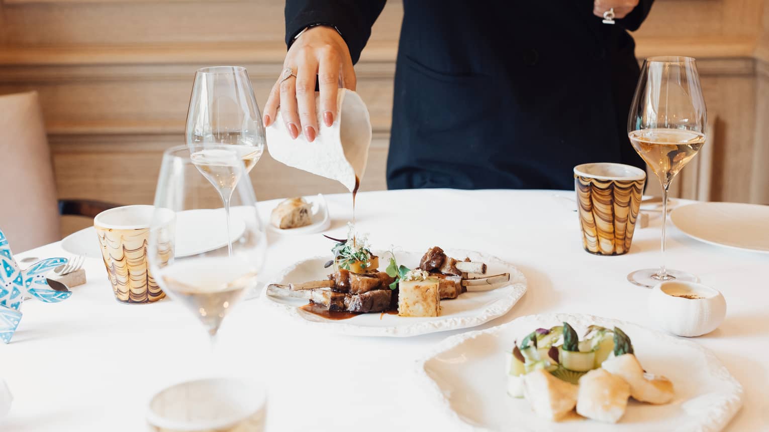 In a well-lit dining space, a person pours a sauce over a plate of food that is set on a round white table set with plates, tumblers, clear stemmed glasses and another serving plate of food items.