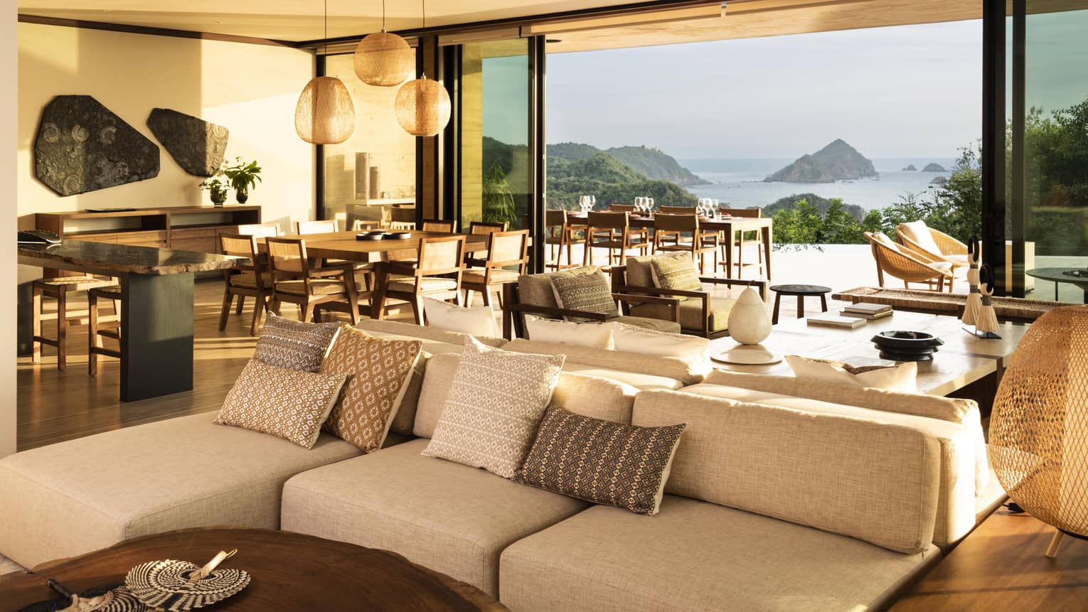 A spacious, open-concept living and dining area in a luxury villa, featuring cozy seating, wooden furnishings and large windows offering stunning views of the ocean and rocky islands