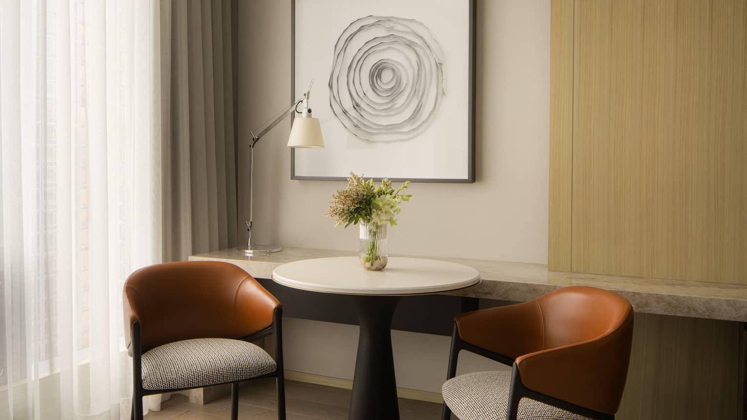 Cozy seating area with two leather chairs, a round table with a vase of flowers, and abstract wall art in a hotel room