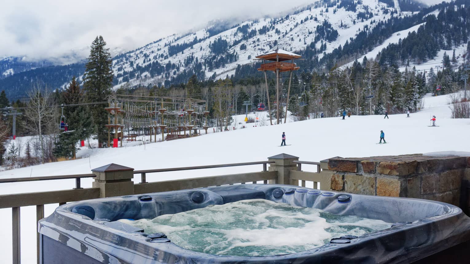Outdoor hot tub with bubbling water overlooking ski slope, rope course and skiers