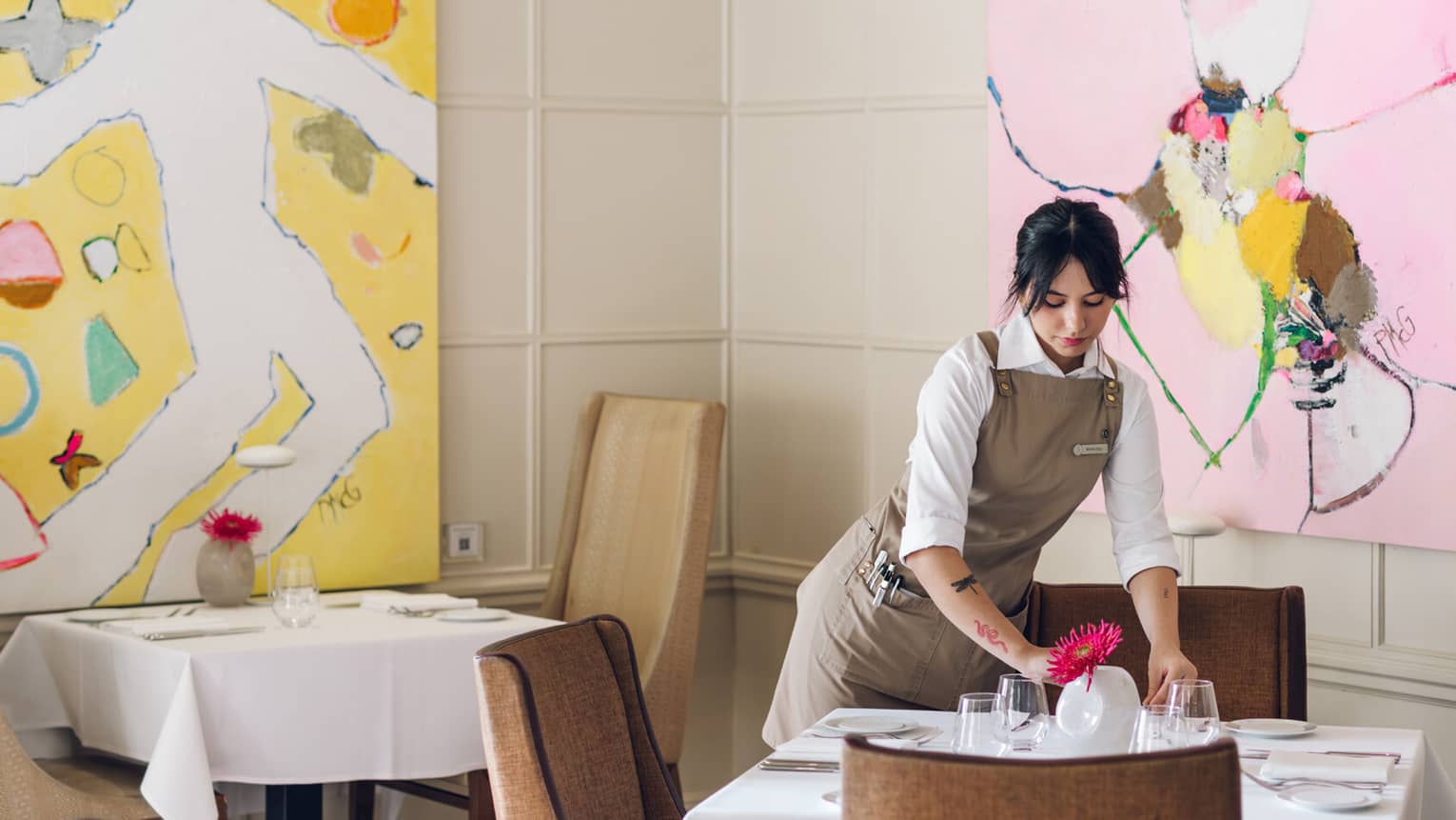 A server standing over a square dining table with a white tablecloth, topped with wine glasses, white dishes and a pink floral arrangement