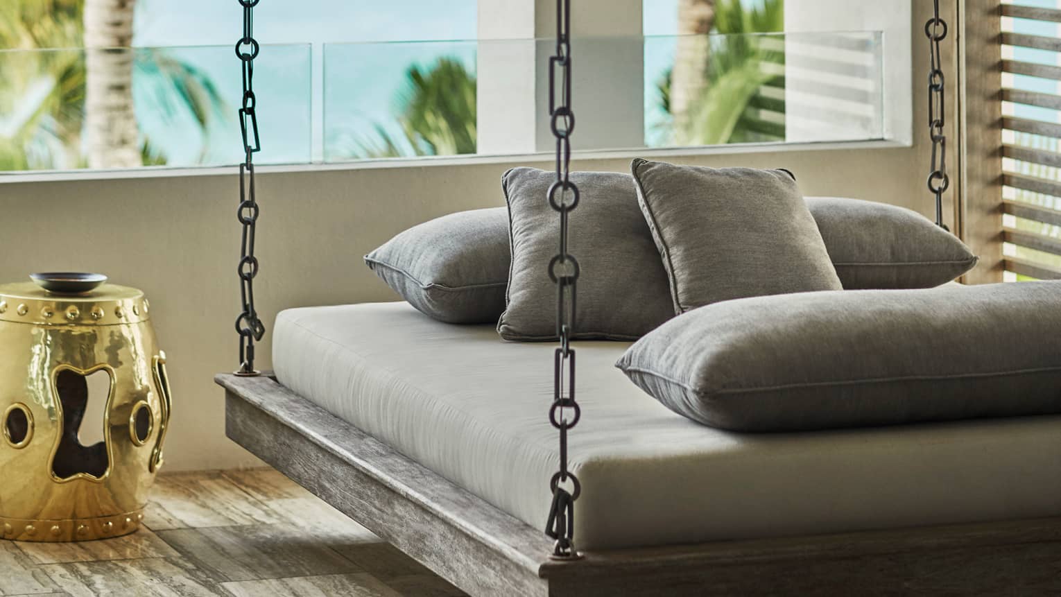 Outdoor hanging daybed with gray cushions and ocean view, beside a gold side table
