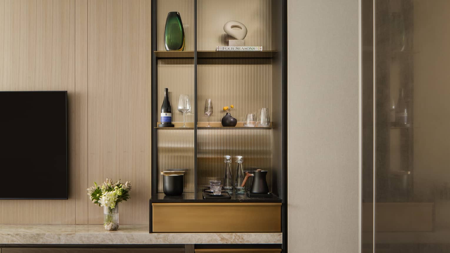 Stylish minibar with glass shelves displaying wine, glasses, and decorative items, next to a TV and vase with fresh flowers