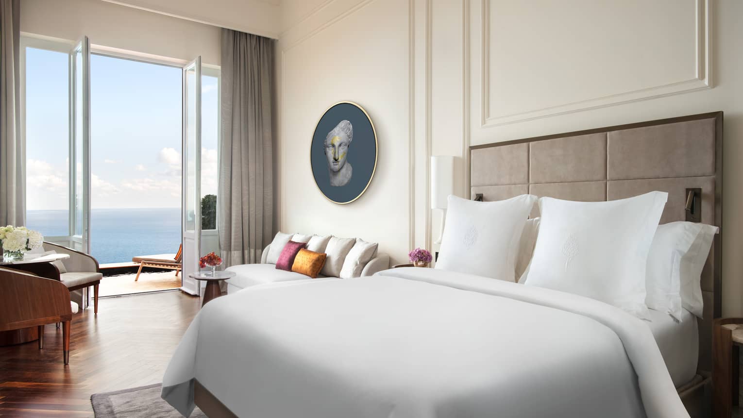 Hotel room with white king bed, white walls with crown molding, wood floors, walk-out sea-view balcony