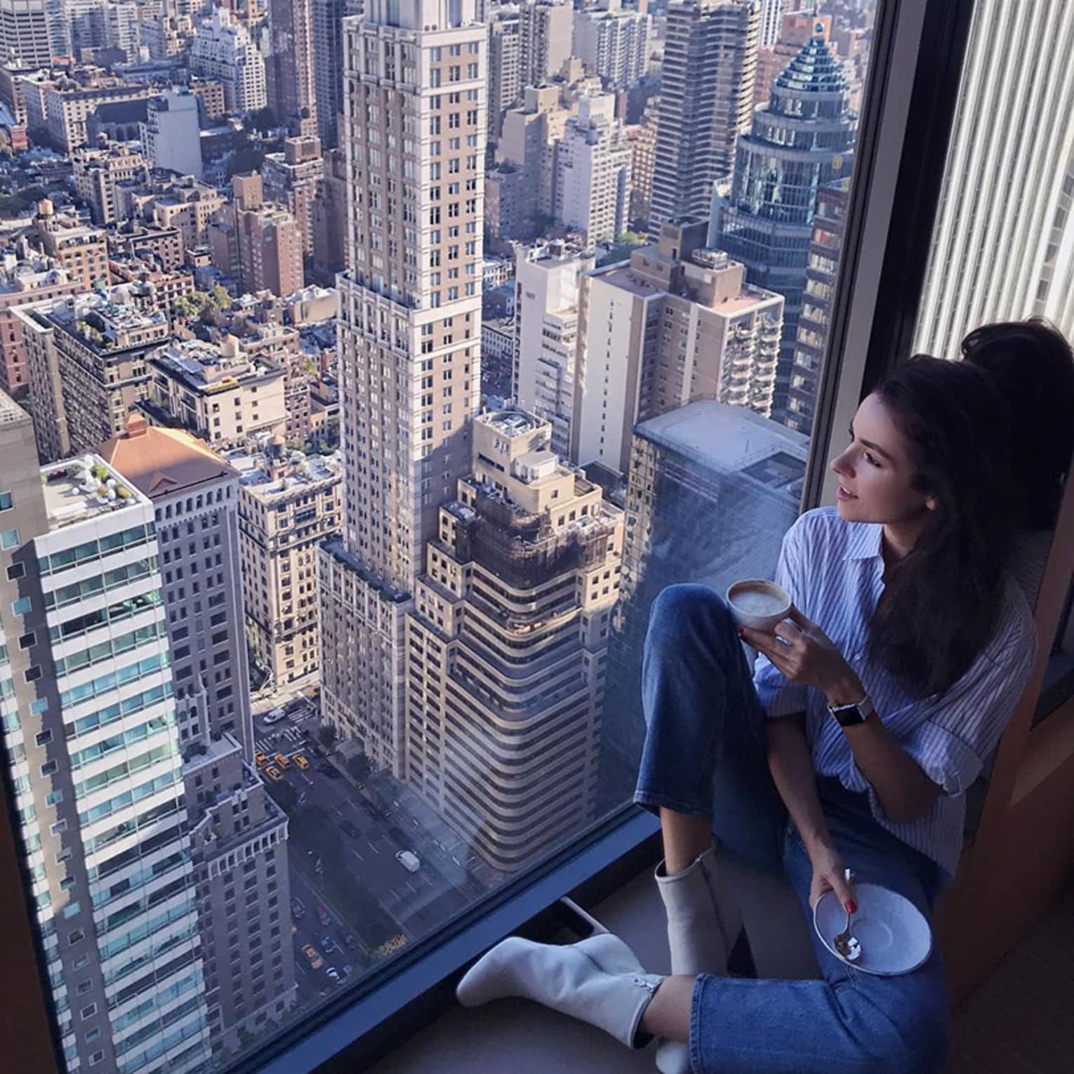 Woman with coffee cup sits in large window overlooking New York City rooftops