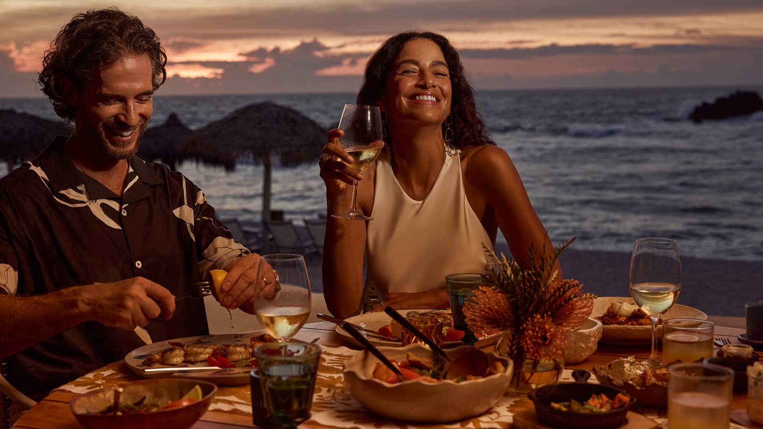 Man and woman laugh while dining at outdoor table filled with food and wine, next to beach and ocean