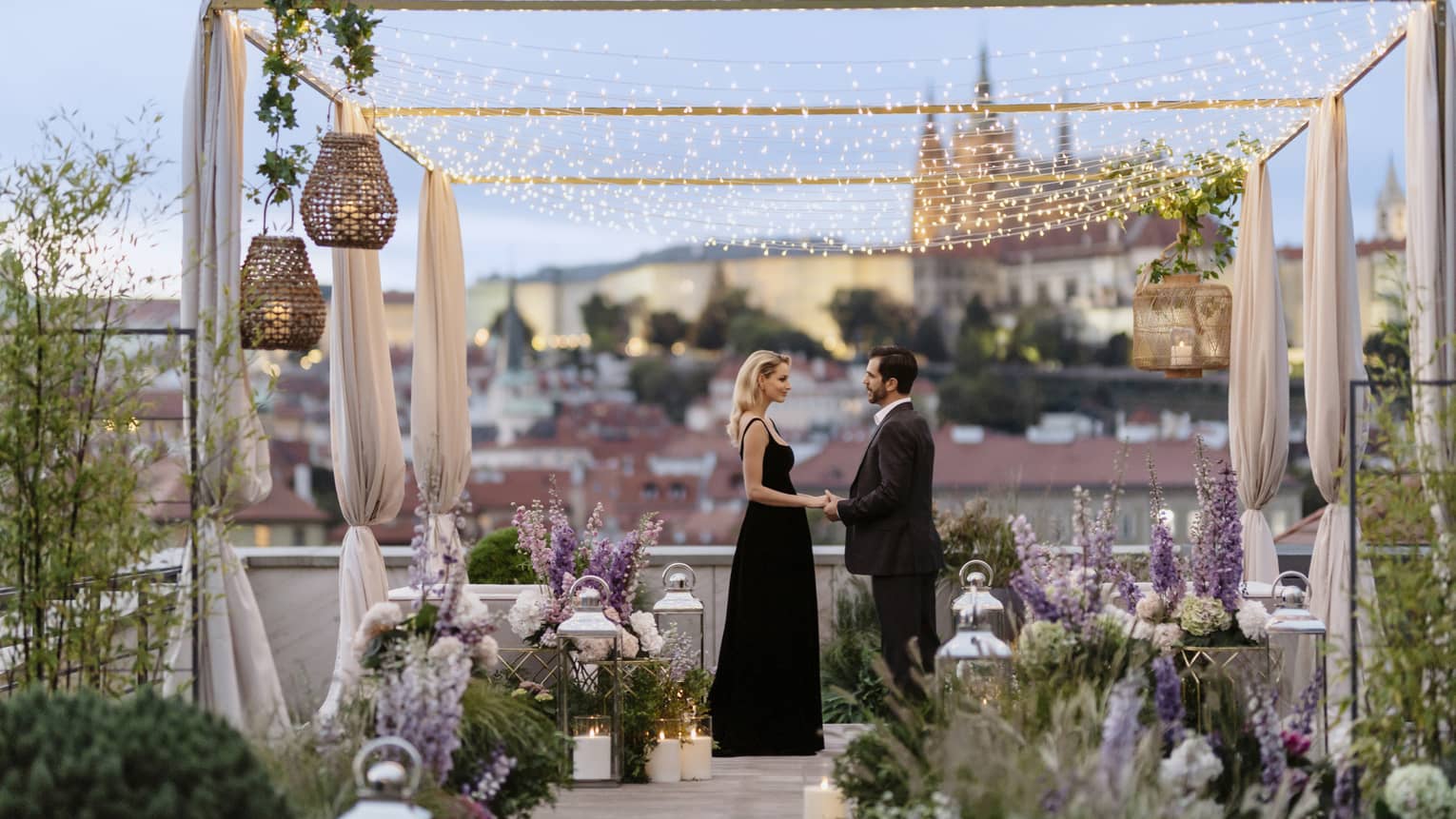 Couple in formalwear holding hands while facing each other on romantically decorated rooftop terrace