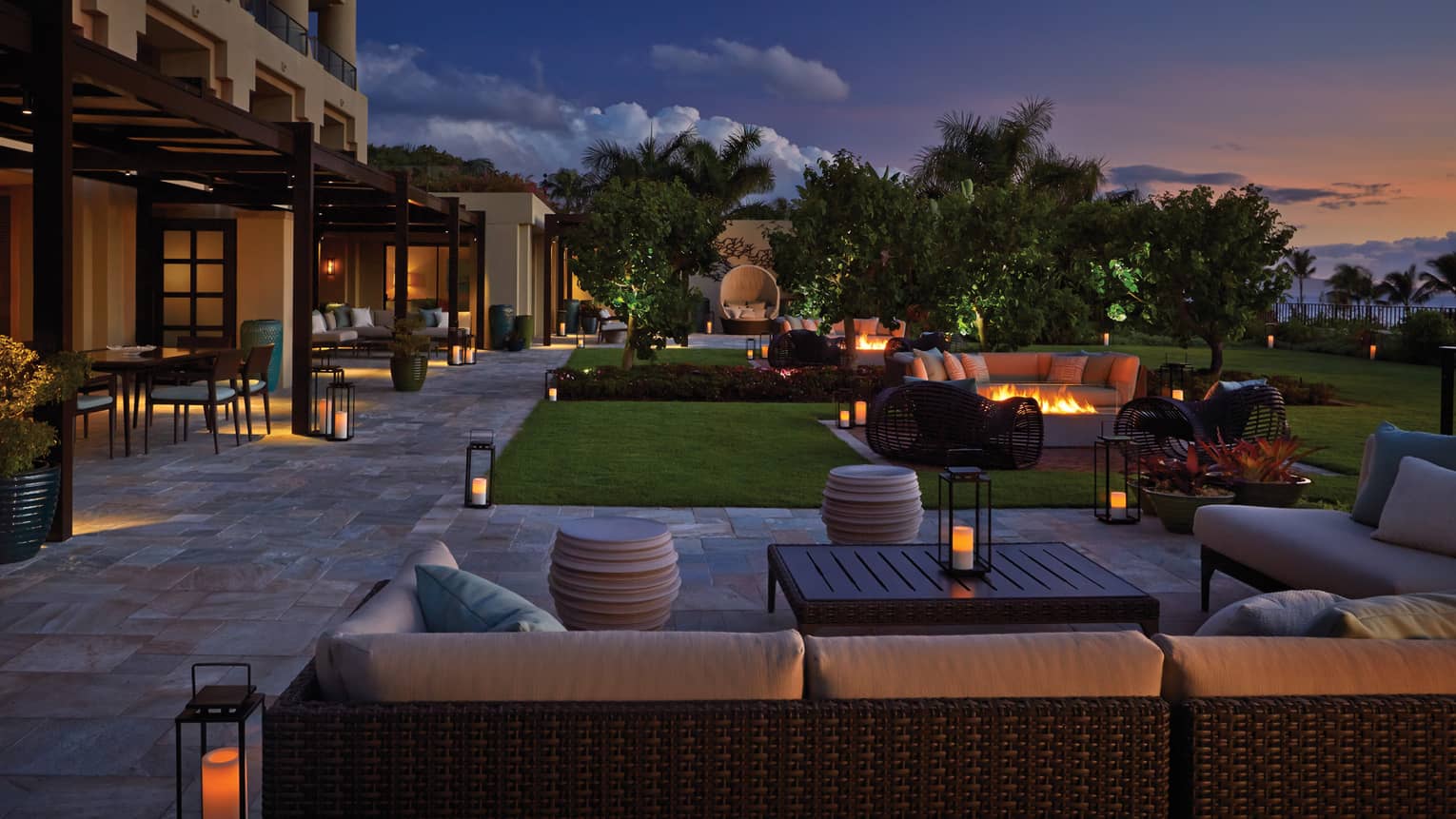Outdoor lounge area at dusk with wicker seating, fire pits, lanterns and garden landscaping