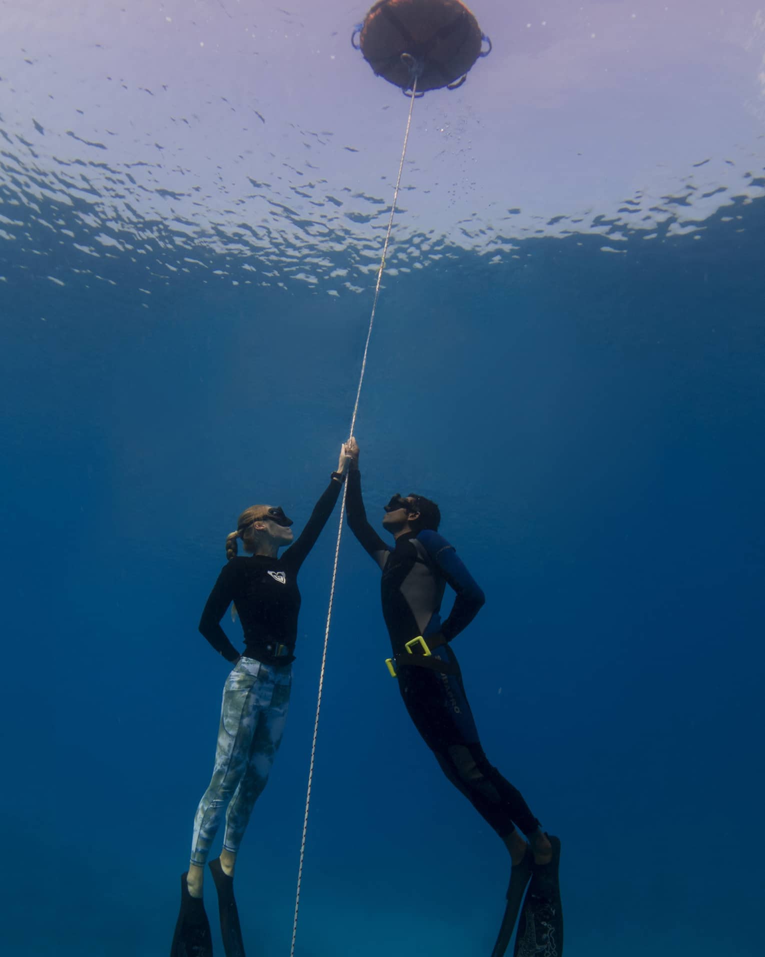 Underwater view of two free-divers, wearing masks and holding onto a rope that attaches to a buoy at the water's surface.