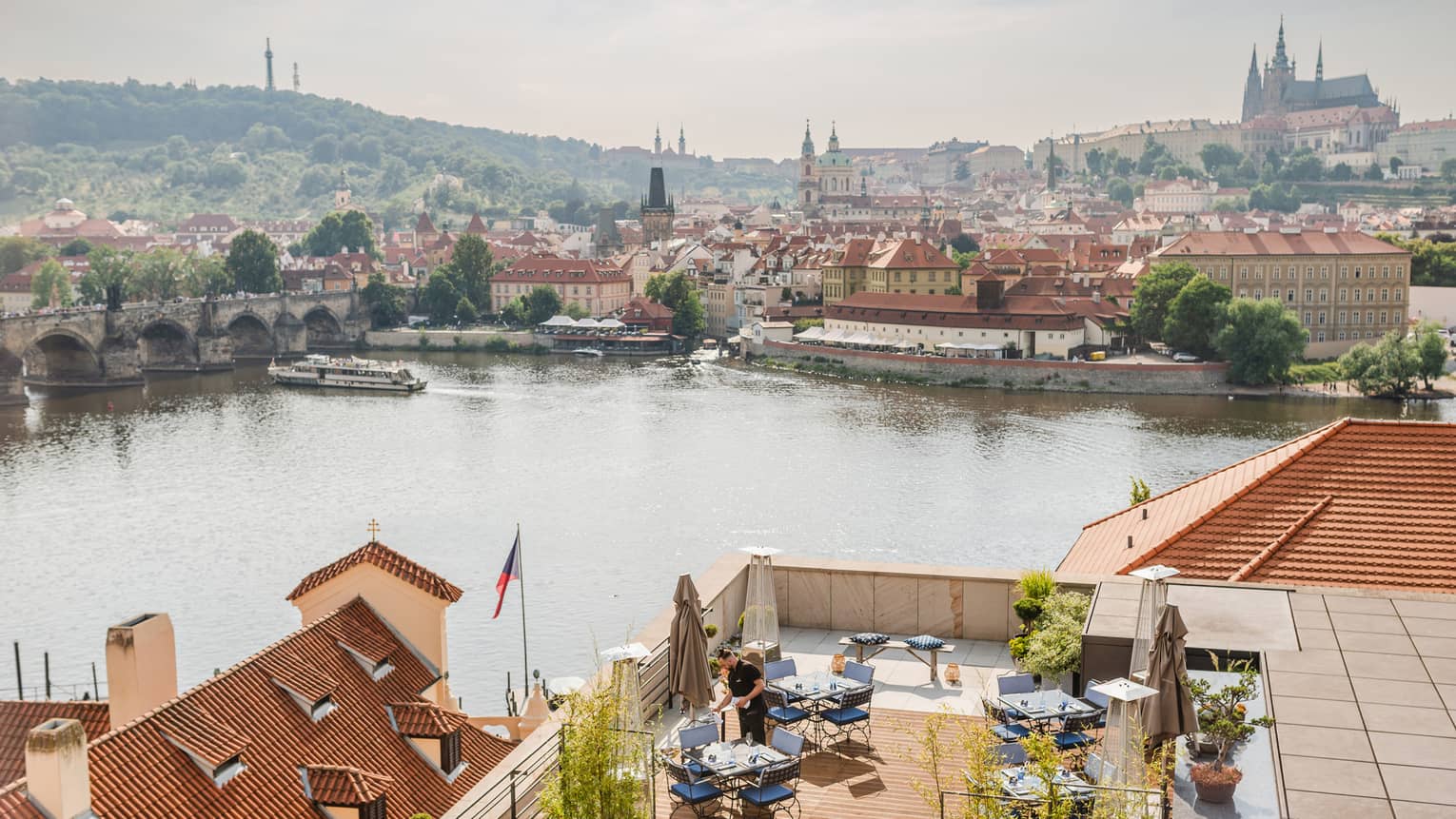 Overhead view of a ferry nearing a medieval stone arch bridge, an ancient castle and cathedral in the distance