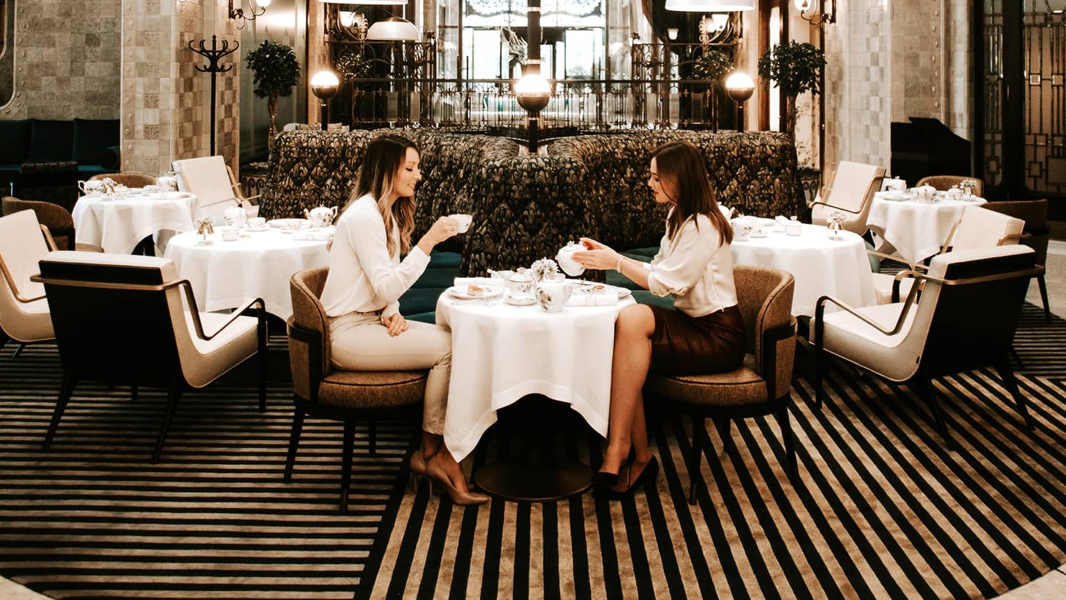 Elegant dining area with a grand chandelier, high arched glass ceiling and modern seating, where two people are enjoying tea at a small table