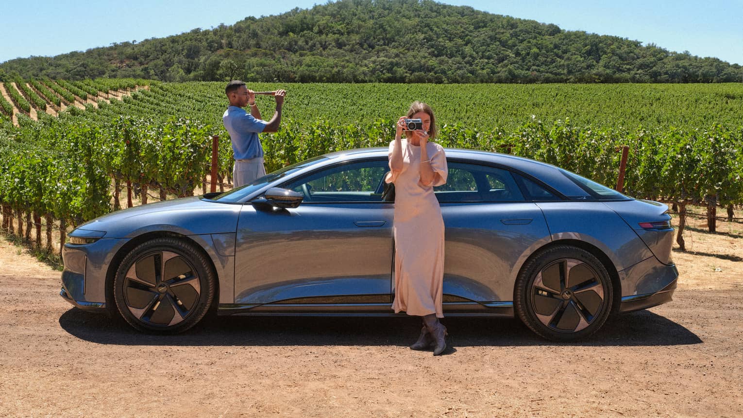 Person wearing long light-pink dress stands leaning against a grey car taking a picture of the surrounding vineyards with their camera while another person wearing a blue shirt and white pants stand on the other side of the car