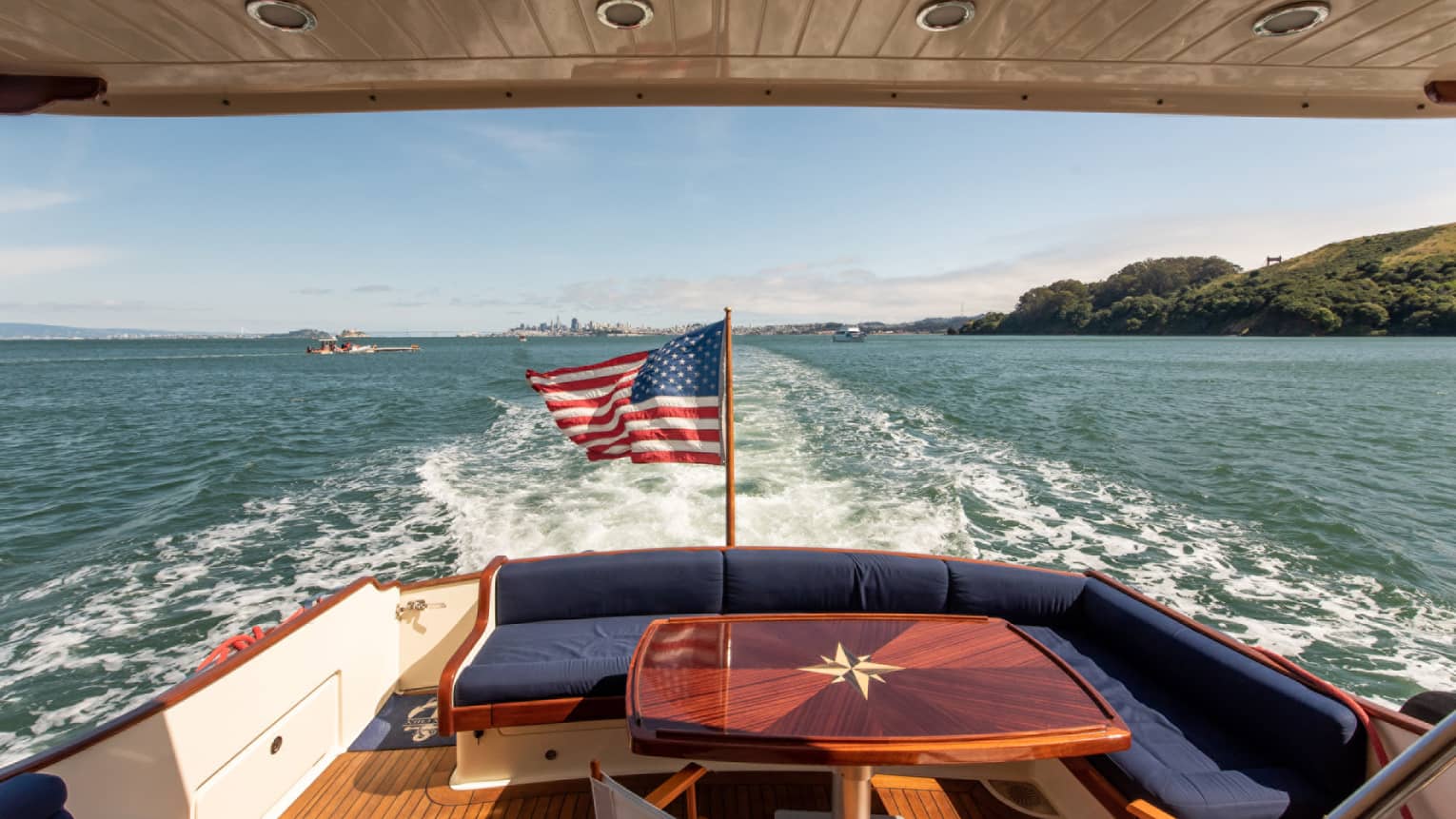 The back of a yacht, with a red table and an American flag, moving along the water.