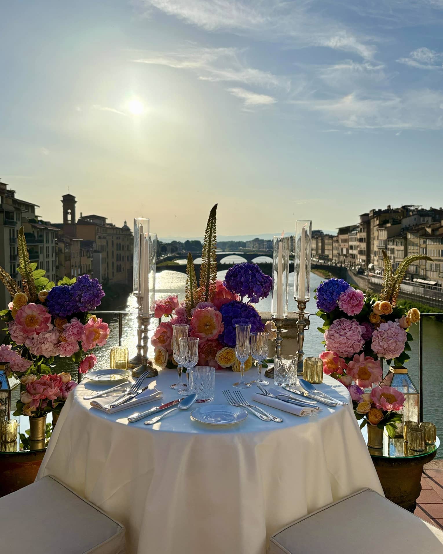 An elegantly set outdoor dining table overlooking a a river, with a sunny sky in the background.