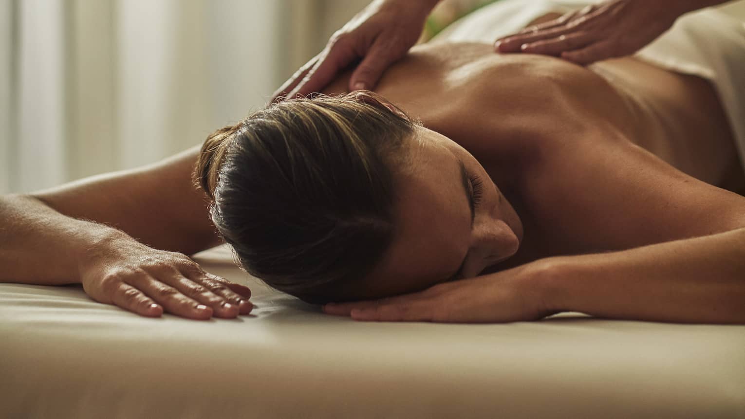 Woman in black uniform massages bare shoulders of woman lying on table face-down with head to side