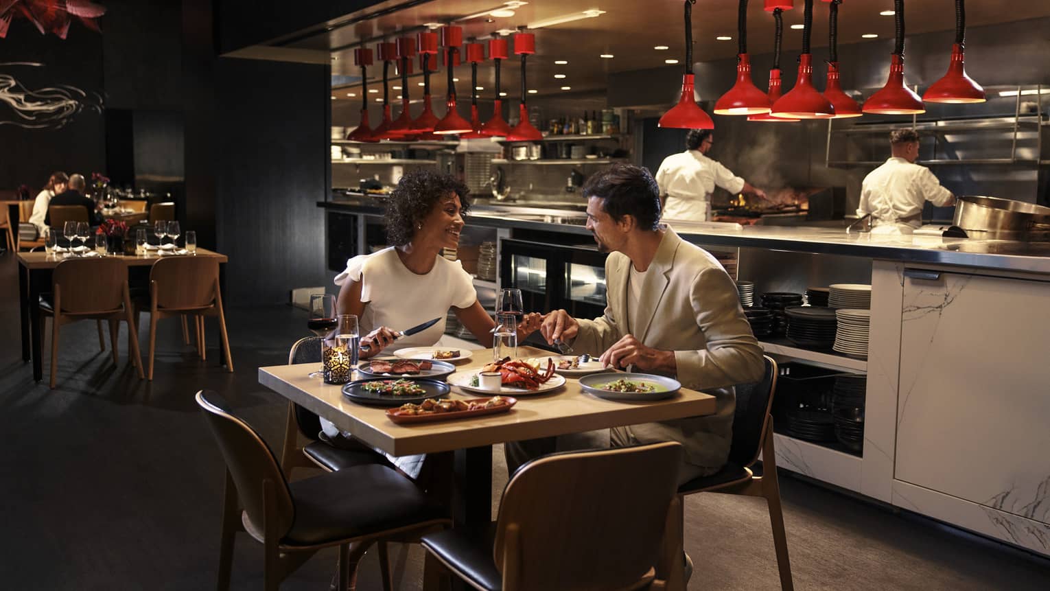 Two people sit at a square dining table in front of an open kitchen with two chefs working behind them