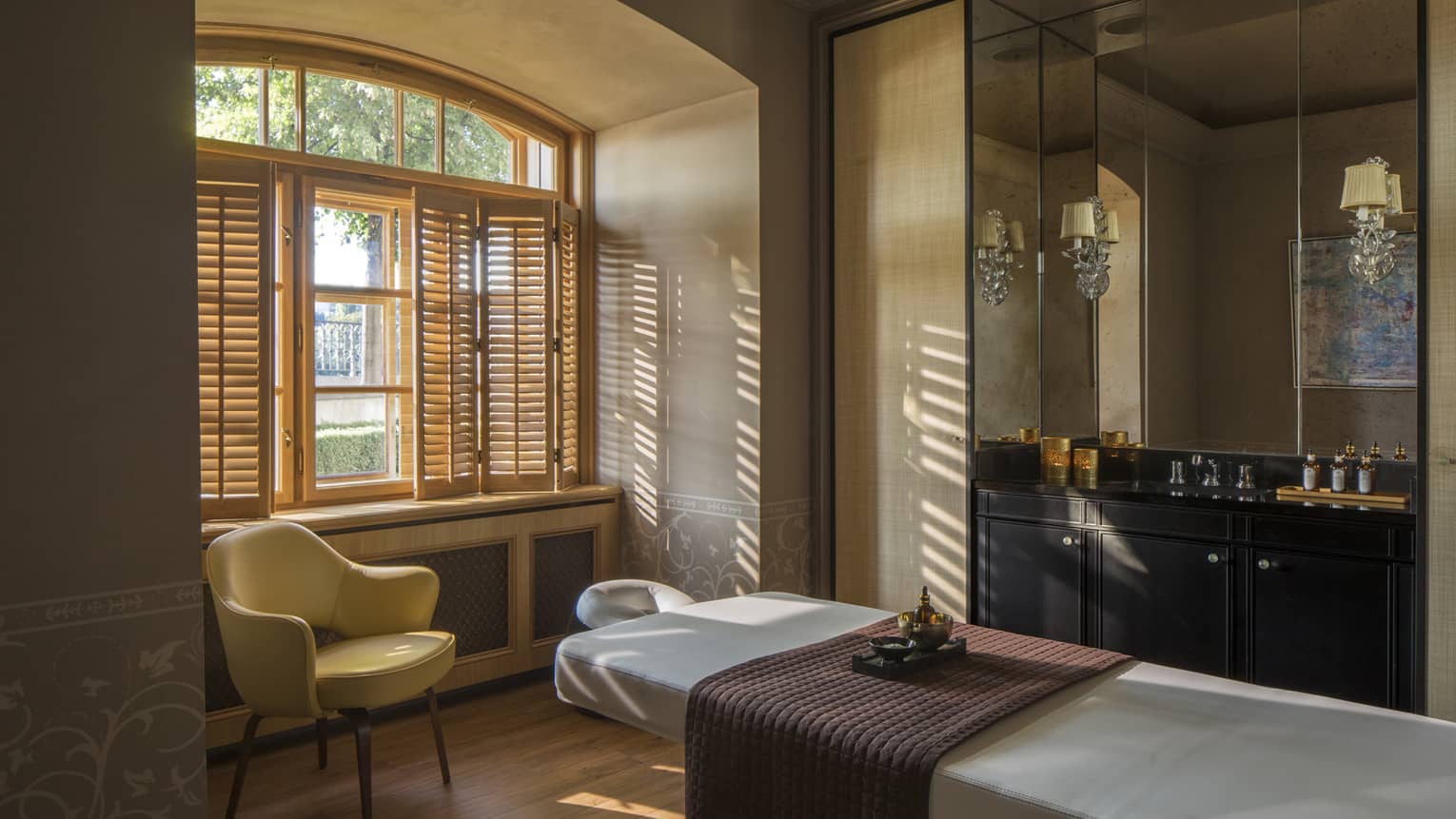 White massage table with quilted brown blanket, mid-century style armchair in front of sunny window, open shutters