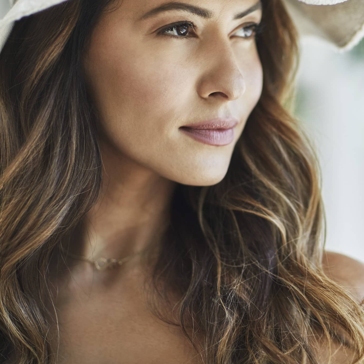 A woman with wavy brown hair and a white floppy hat looks off into the distance