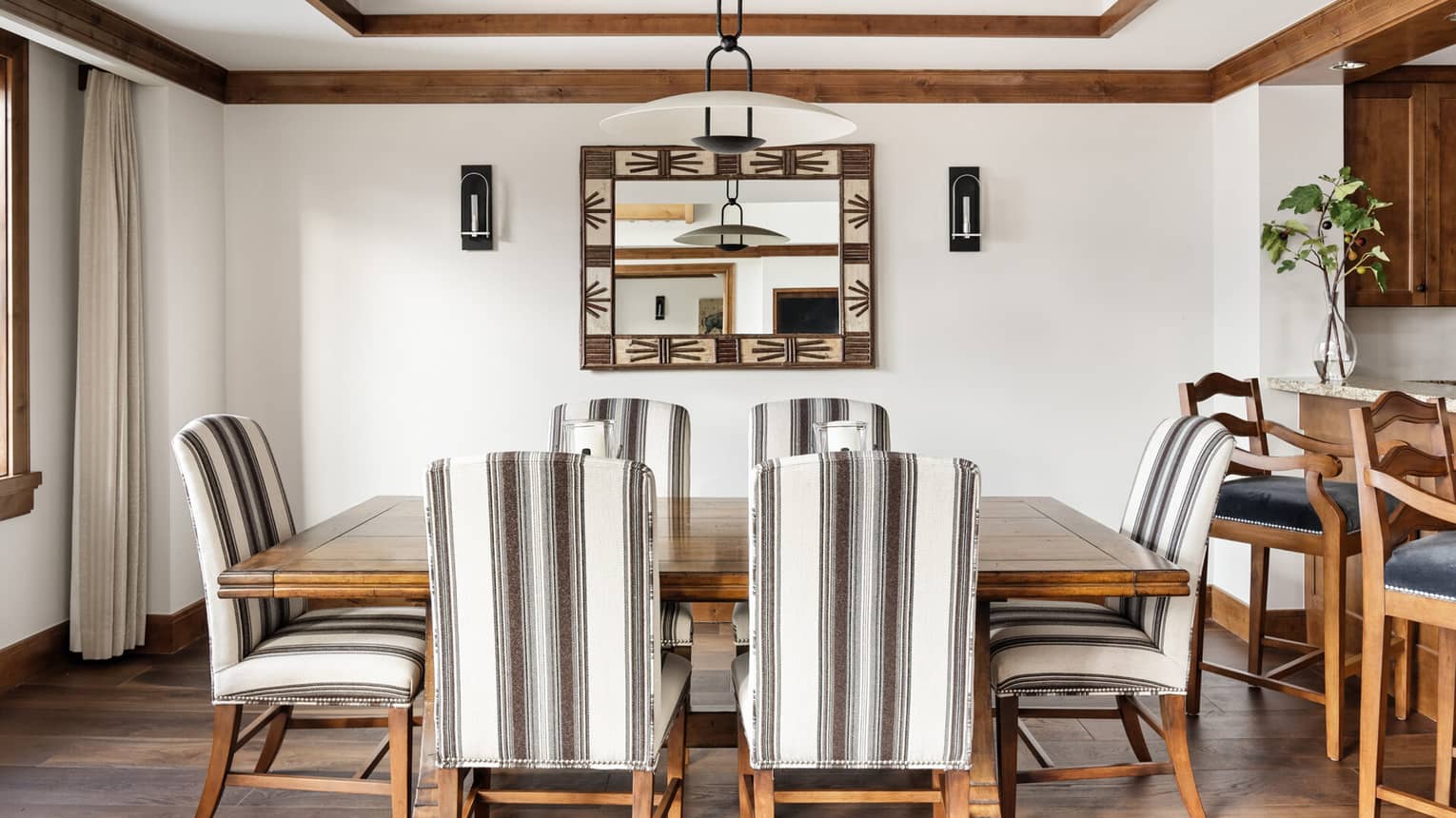 Dining room with a wooden table surrounded by six striped upholstered chairs and decorative mirror flanked by two wall sconces