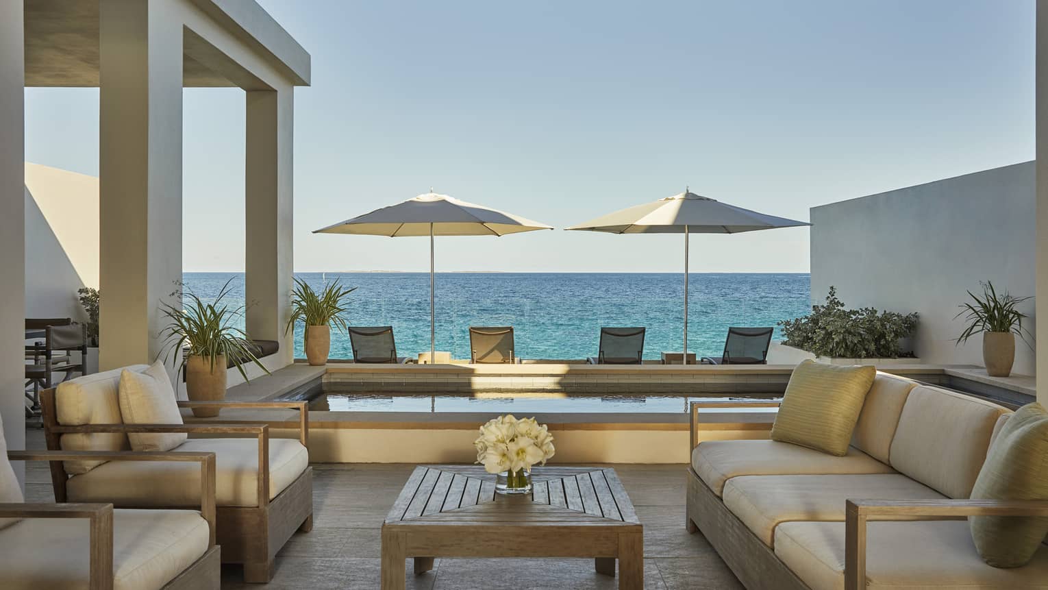 Outdoor terrace with cushioned seating, a wooden table, two umbrellas and ocean views beyond a private pool