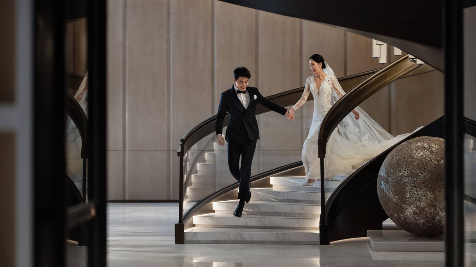 A smiling bride and groom walk hand in hand down the stairs at Four Seasons Hotel Dalian.