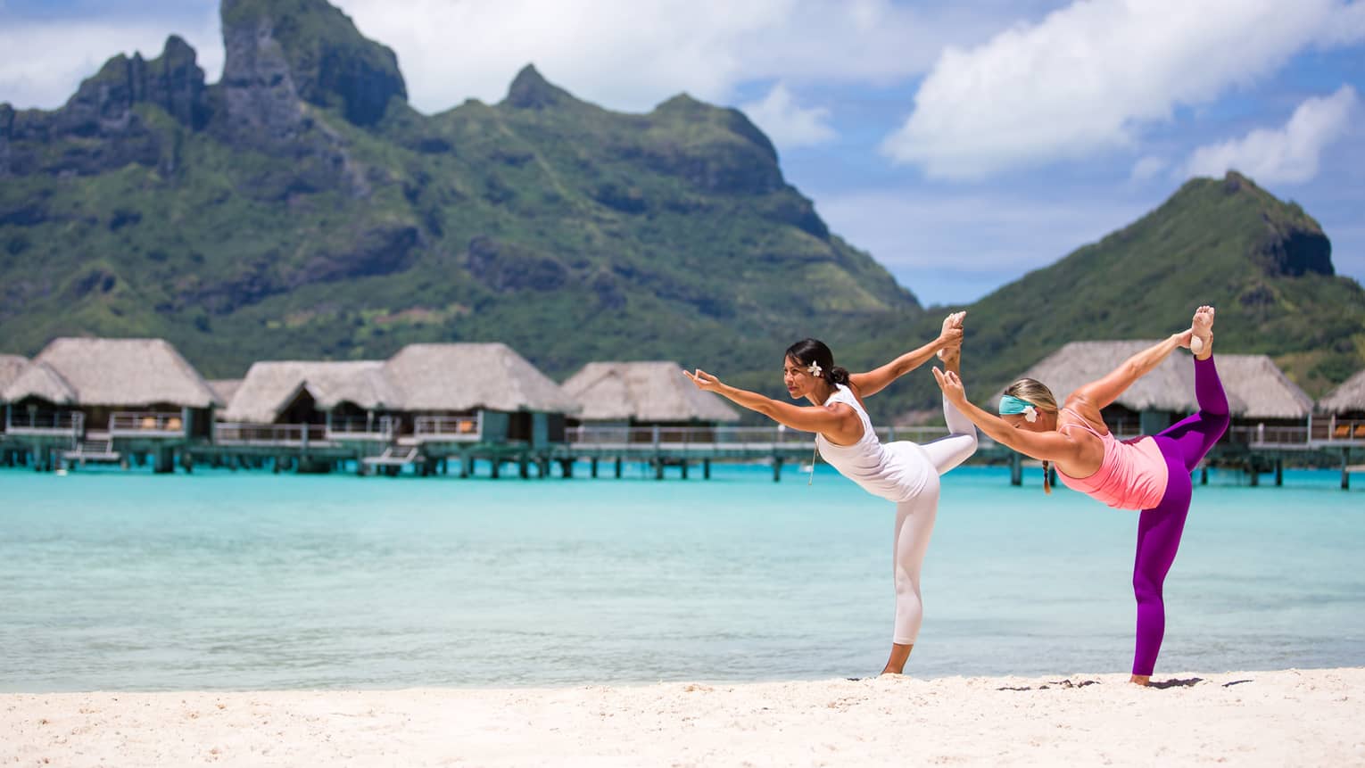 Two people practicing yoga on a tropical beach with overwater bungalows and mountains in the background