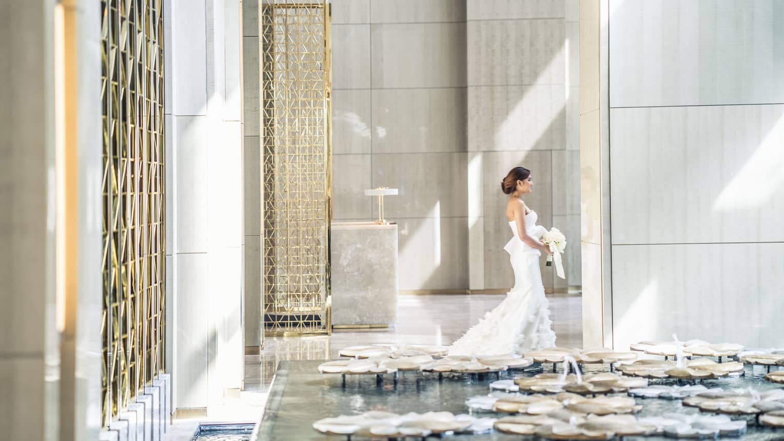 Sun rays illuminates a bride as she walks through a white walled lobby carrying her bouquet of white flowers